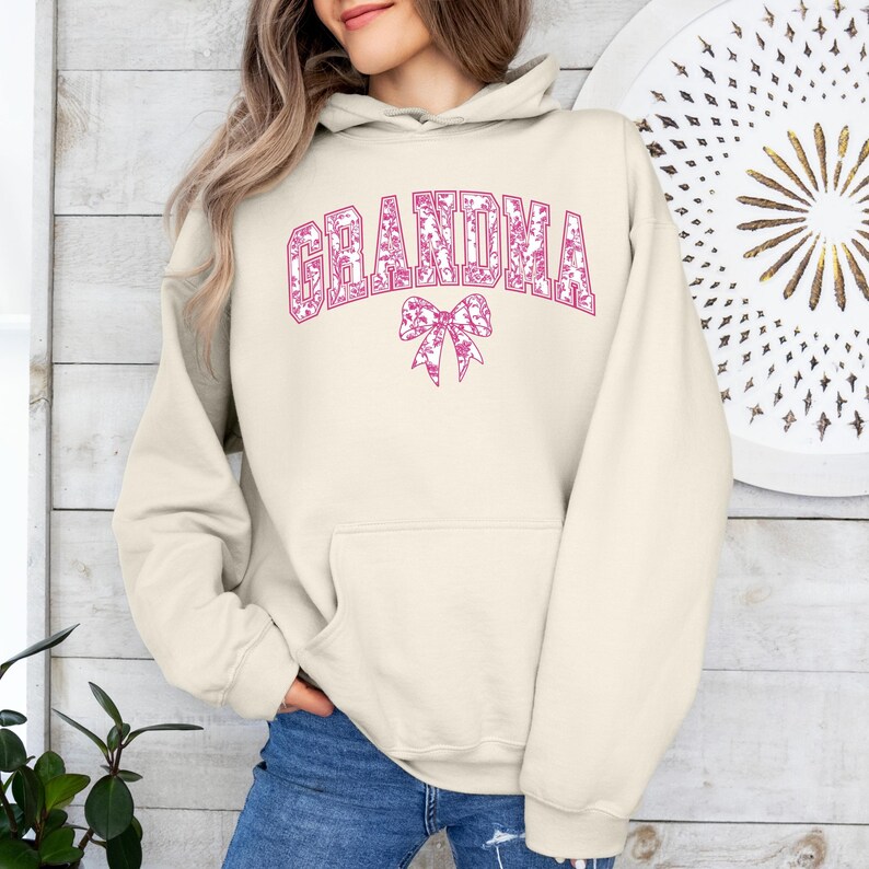 An olive green sweatshirt with "GRANDMA" printed in bold pink floral letters, accented by a matching pink bow. Worn by a woman styled with ripped denim jeans in a cozy, modern setting. A perfect gift for fashionable grandmothers.