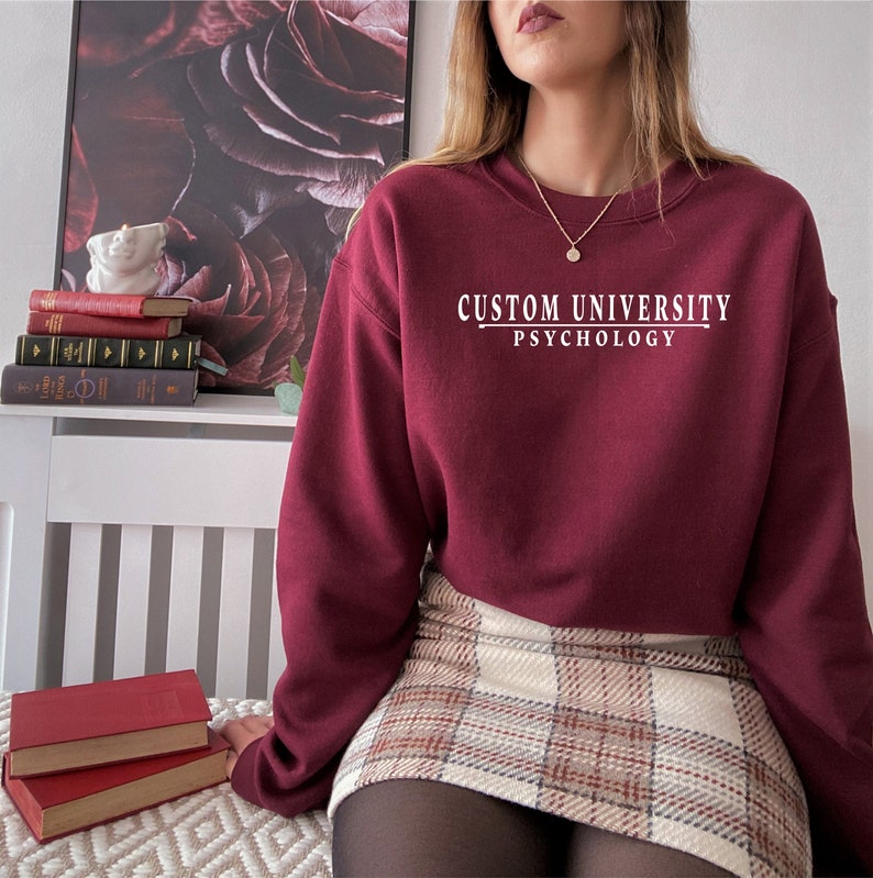 May include: A maroon sweatshirt with the text "CUSTOM UNIVERSITY PSYCHOLOGY" in white lettering. The sweatshirt is worn by a person wearing a plaid skirt and black tights.