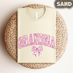 A beige t-shirt with the word "GRANDMA" printed in pink letters, along with a pink bow design, is displayed on a woven basket.