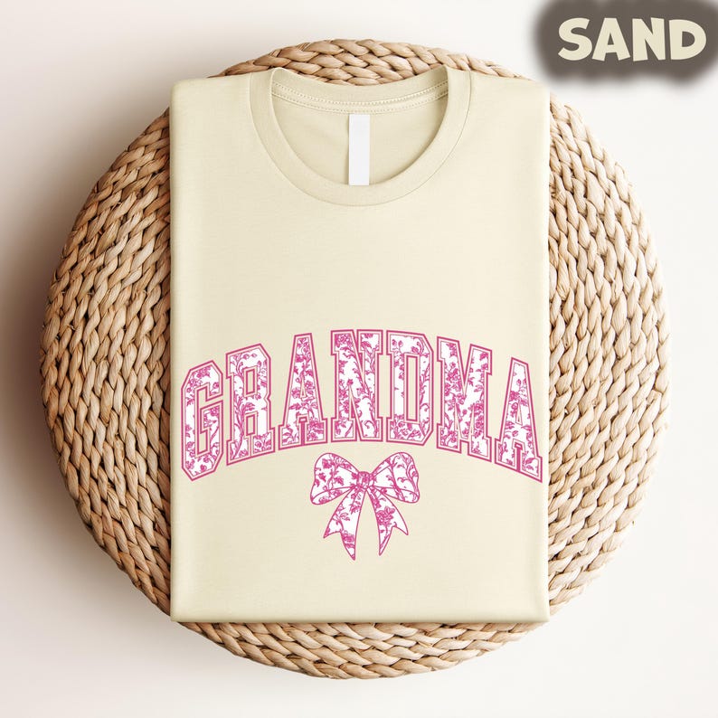 A beige t-shirt with the word "GRANDMA" printed in pink letters, along with a pink bow design, is displayed on a woven basket.