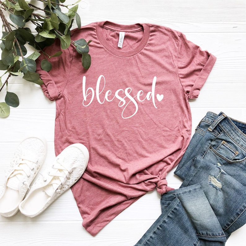 May include: A dusty rose colored t-shirt with the word "blessed" in white script with a small white heart next to it. The shirt is laid flat on a white surface with a pair of white lace-up shoes and a pair of blue jeans.