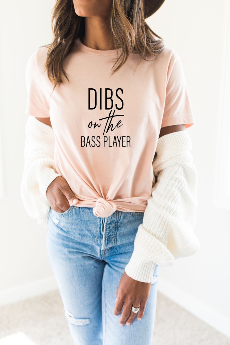 May include: A light pink t-shirt with the text "DIBS on the BASS PLAYER" in black. The shirt is knotted at the waist and paired with blue jeans. A white sweater is draped over the shoulders.