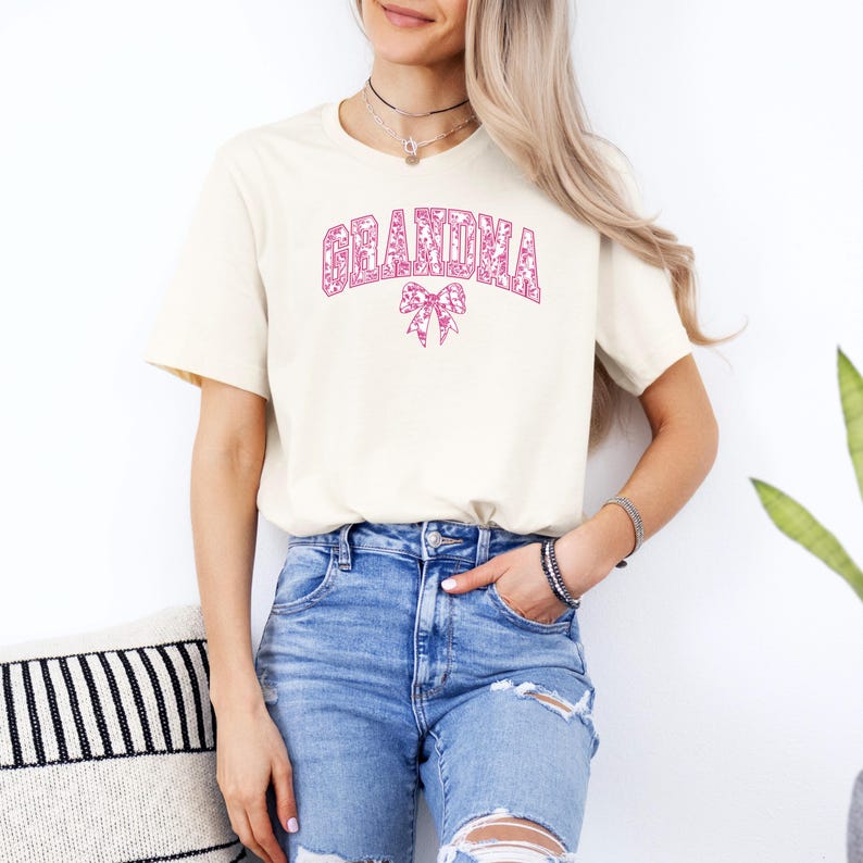 An olive green sweatshirt with "GRANDMA" printed in bold pink floral letters, accented by a matching pink bow. Worn by a woman styled with ripped denim jeans in a cozy, modern setting. A perfect gift for fashionable grandmothers.