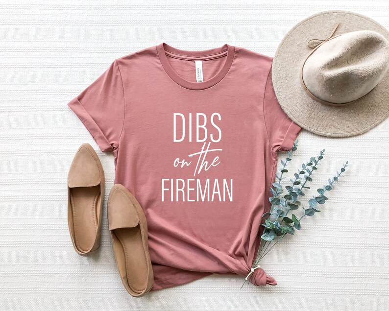 May include: Dusty rose t-shirt with the text "DIBS on the FIREMAN" in white. The shirt is styled with a beige hat, beige loafers, and a sprig of eucalyptus. The t-shirt is made of a soft material.