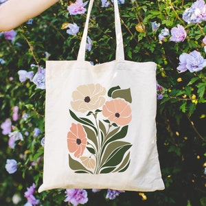 May include: A white canvas tote bag with a floral design. The flowers are in shades of pink, peach, and cream. The leaves are green.