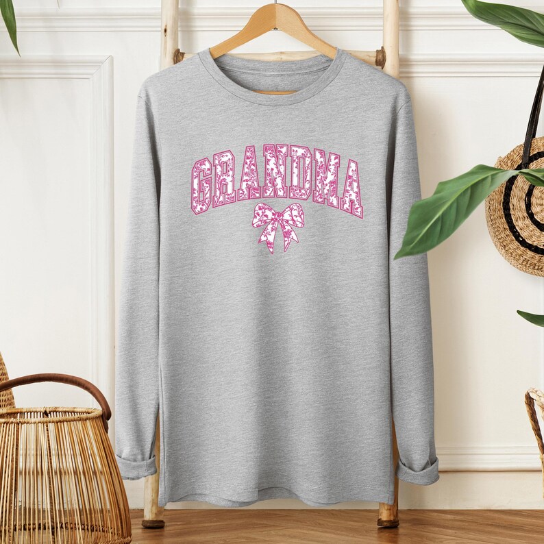 An olive green sweatshirt with "GRANDMA" printed in bold pink floral letters, accented by a matching pink bow. Worn by a woman styled with ripped denim jeans in a cozy, modern setting. A perfect gift for fashionable grandmothers.