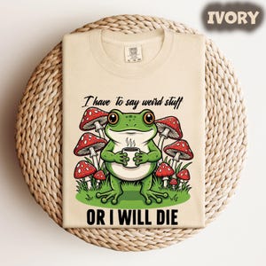 May include: Ivory-colored t-shirt featuring a green frog holding a coffee cup, surrounded by red and white mushrooms. The text "I have to say weird stuff OR I WILL DIE" is printed below the frog. The shirt is folded on a woven surface.