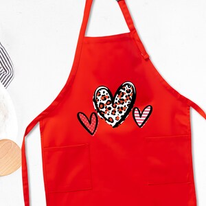 May include: A bright red apron featuring three heart designs. The central heart has a black outline with a leopard print pattern in red and white. The other two hearts are outlined in black with red and white accents. The apron has two front pockets.