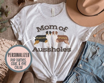 Puede incluir: Una camiseta gris con el texto "Mom of Aussholes" y dos perros pastores australianos de dibujos animados con los nombres "Popcorn" y "Whiskey" impresos en ella.