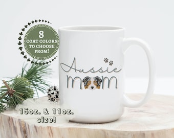 May include: White ceramic mug with the text "Aussie Mom" in blue and brown lettering. A cartoon Australian Shepherd dog is in the center of the text. The mug is available in 15 oz. and 11 oz. sizes.  The text "8 Coat Colors to Choose From!" is in a green circle with white leaves.