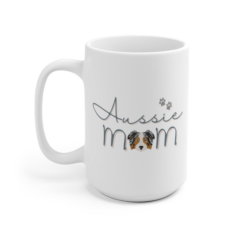 Op de afbeelding: Witte keramische mok met een blauw en roze ontwerp met de tekst "Aussie Mom" en een cartoon Australian Shepherd-hond in het midden.