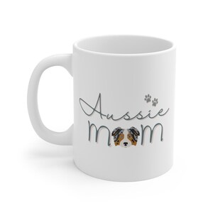 Op de afbeelding: Witte keramische mok met een blauw en grijs ontwerp met de tekst "Aussie Mom" en een cartoon Australian Shepherd-hond in het midden.