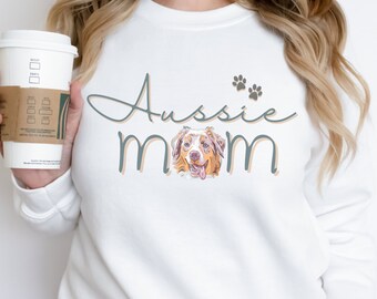 Puede incluir: Sudadera blanca con la frase "Aussie mom" en fuente cursiva, con un gráfico de un perro pastor australiano. Dos huellas de patas están encima del texto. La sudadera es de estilo casual.