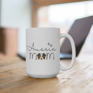 Op de afbeelding: Witte keramische koffiemok met een blauwe en grijze afbeelding van een Australian Shepherd hond en de tekst "Aussie mom" met poot afdrukken.
