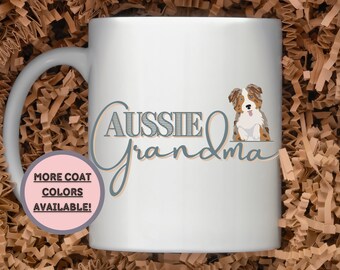 May include: White ceramic mug with a blue and brown Australian Shepherd dog illustration and the text "Aussie Grandma" in blue and peach. A pink circle with the text "More Coat Colors Available!" is in the lower left corner.