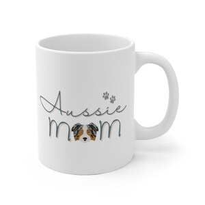 Op de afbeelding: Witte keramische koffiemok met een blauw en grijs ontwerp met de tekst "Aussie Mom" met een pootprint en een cartoon Australian Shepherd-hond.