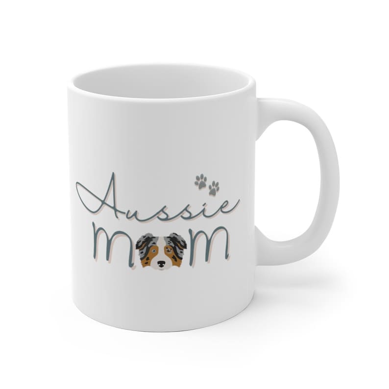 Op de afbeelding: Witte keramische koffiemok met een blauw en grijs ontwerp met de tekst "Aussie Mom" met een pootprint en een cartoon Australian Shepherd-hond.