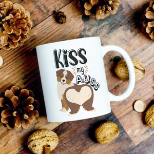 May include: White ceramic mug with a brown and white cartoon Australian Shepherd dog with the text "Kiss my Aus" in black lettering.