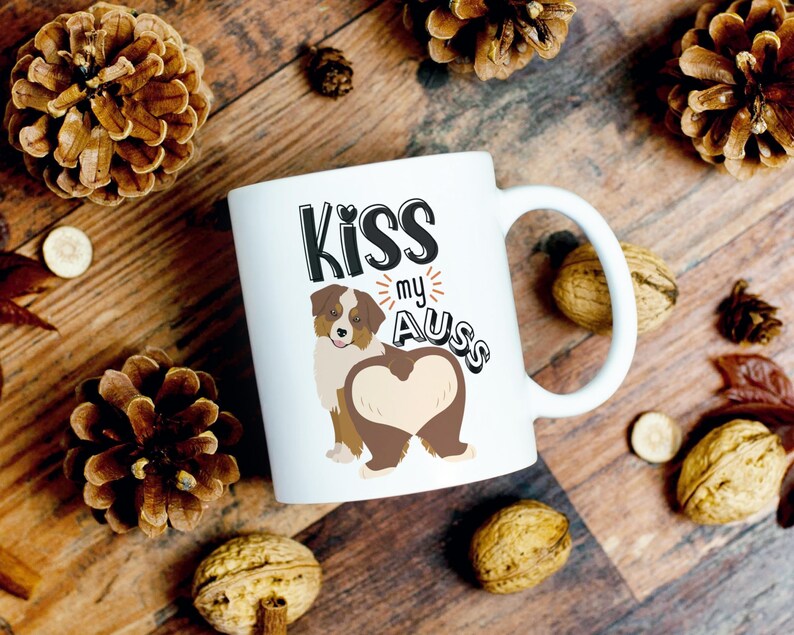 May include: White ceramic mug with a brown and white cartoon Australian Shepherd dog with the text "Kiss my Aus" in black lettering.