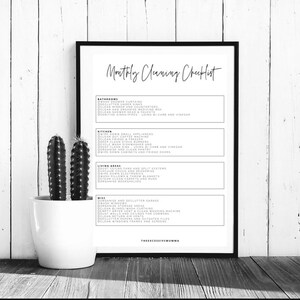 May include: A framed black and white print titled "Monthly Cleaning Checklist". The checklist includes tasks for bathrooms, kitchen, living areas, and miscellaneous areas. A potted cactus sits to the left of the frame on a wooden surface.