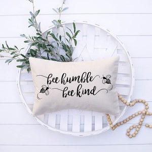 May include: A white pillow with black text that reads "bee humble bee kind". Two black and yellow bees are on either side of the text. The pillow is sitting on a white wicker basket.