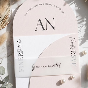 Könnte beinhalten: Eine bogenförmige, blassrosa Einladungskarte mit den schwarzen Buchstaben AN und dem Text We can't wait to celebrate with you. Ein halbtransparentes Pergamentband umschließt die Karte, bedruckt mit Finer Details, Kindly RSVP und You are invited.