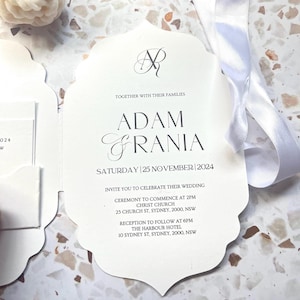 Puede incluir: Una invitación de boda blanca con un borde decorativo, con los nombres "Adam & Grania" y la fecha "Sábado 25 de noviembre de 2024." Incluye detalles de la ceremonia y la recepción. Una cinta blanca está atada.