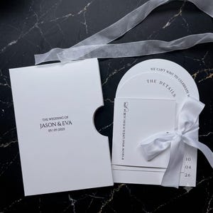May include: A white wedding invitation suite featuring a rectangular card with a thumb notch, a semi-circular details card, and a square card with the text IF I KNOW WHAT LOVE IS IT IS BECAUSE OF YOU, all tied together with a white satin ribbon.