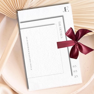 May include: A white card with the text "THE DETAILS" and "JE" in black lettering. A burgundy ribbon with a bow is tied around the card. The date "03 05 25" is written in black on the card.