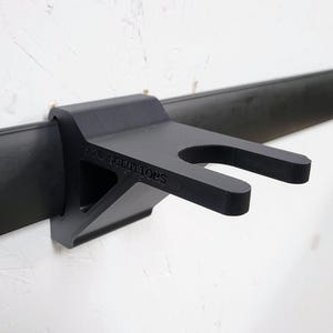 Peut inclure: Support noir en forme de U pour maintenir un objet, monté sur une barre noire. Le support est fixé à un mur blanc. Le texte "V2C SOLUTIONS" est imprimé sur le support.