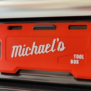 Puede incluir: Una placa rectangular de plástico naranja con el texto en relieve blanco Michael's y TOOL BOX. La placa tiene un clip horizontal negro en la parte superior central y dos recortes rectangulares a cada lado para su montaje.