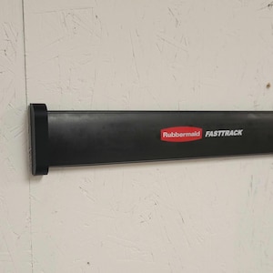 Op de afbeelding: Een zwarte Rubbermaid FastTrack wandmontagerail met een glad, horizontaal ontwerp en een rood-wit logo op het oppervlak. De rail is bevestigd aan een lichtgekleurde, getextureerde muur en biedt een stevige basis voor opslag.
