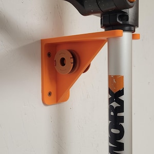 May include: An orange 3D-printed bracket mounted on a wall, designed to hold a WORX brand tool. The bracket has a triangular shape with a circular holder and is secured with screws. The tool itself is black and silver.