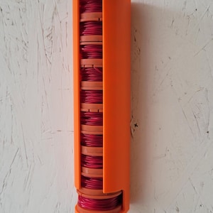 May include: A vertical orange plastic storage dispenser holding a stack of ten circular spools wound with bright pink wire. The dispenser features a long open slot along the front, allowing the individual spools to be seen and accessed easily.