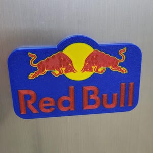 May include: A blue and yellow Red Bull logo with two red bulls facing each other. The words "Red Bull" are in red, and the logo is set against a silver background.