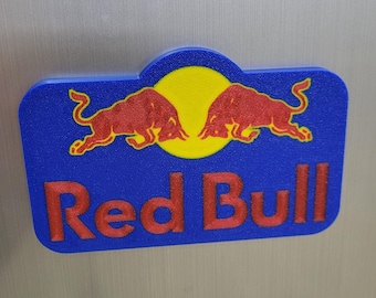 Op de afbeelding: Een blauw en geel Red Bull-logo met twee rode stieren tegenover elkaar. De tekst "Red Bull" is rood. Het logo staat tegen een zilveren achtergrond.