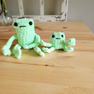 May include: Two crocheted green frog plushies, one larger than the other, sitting on a wooden surface.