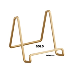 May include: Gold metal display stand for showcasing small items like photos, artwork, or figurines. The stand has a simple, triangular design.