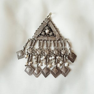 Puede incluir: Un colgante de plata con una parte superior triangular y una franja de pequeños cuadrados de plata y perlas blancas.