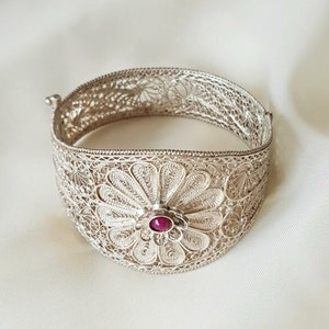 Könnte beinhalten: Ein silbernes Filigranarmband mit einem roten Edelstein in der Mitte eines Blumenmusters.