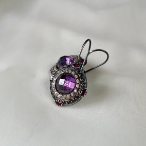Puede incluir: Un par de pendientes de plata con una piedra preciosa p&uacute;rpura rodeada de piedras blancas m&aacute;s peque&ntilde;as y un borde de piedras p&uacute;rpuras m&aacute;s peque&ntilde;as.