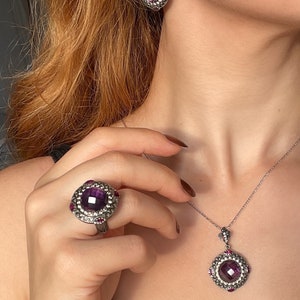Puede incluir: Un conjunto de collar y pendientes de plata con una piedra preciosa p&uacute;rpura rodeada de peque&ntilde;as piedras preciosas rosas. El collar tiene un colgante de dise&ntilde;o cuadrado.