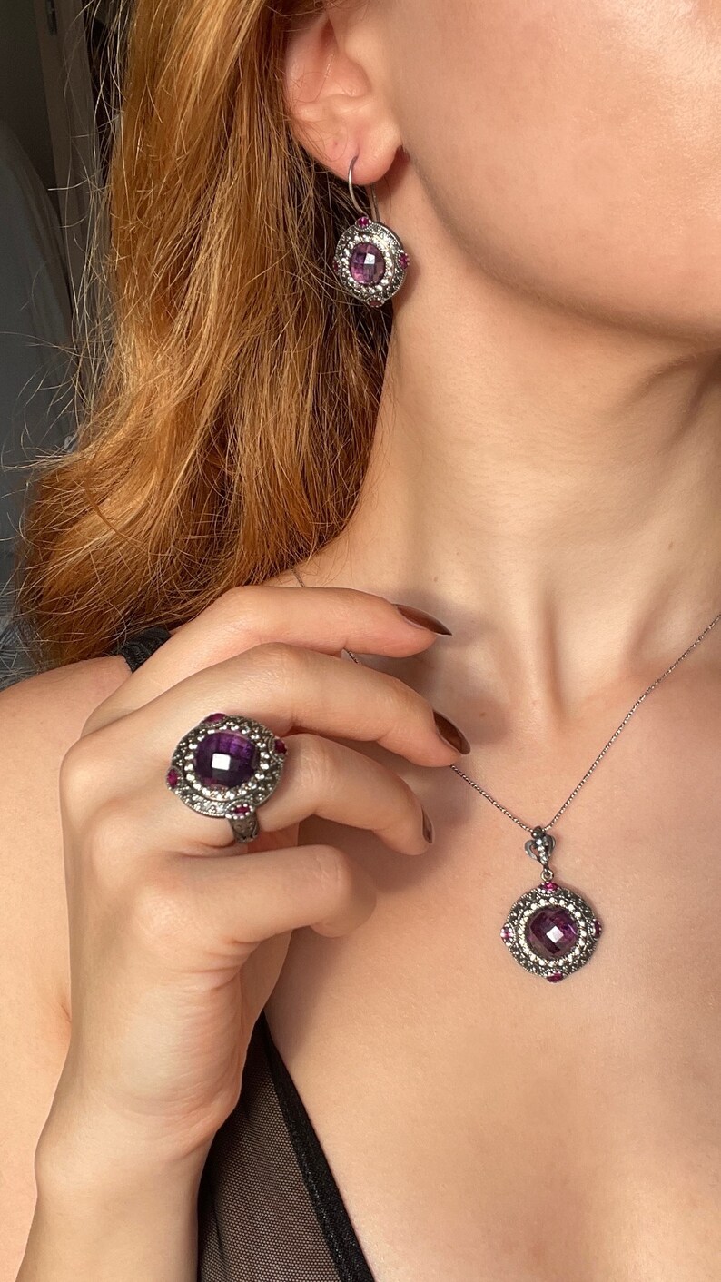 Puede incluir: Un conjunto de collar y pendientes de plata con una piedra preciosa p&uacute;rpura rodeada de peque&ntilde;as piedras preciosas rosas. El collar tiene un colgante de dise&ntilde;o cuadrado.