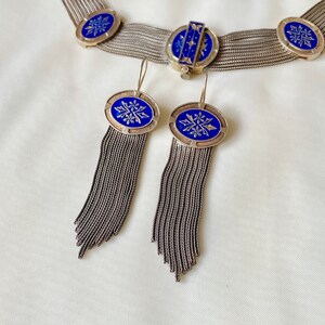 K&ouml;nnte beinhalten: Silberfarbenes Schmuckset mit Halskette und Ohrringen. Die Halskette und die Ohrringe haben ovale blaue Emaille-Akzente mit floralen Designs. Die Ohrringe haben lange, baumelnde Fransen. Das Schmuckst&uuml;ck wird auf einer wei&szlig;en Oberfl&auml;che pr&auml;sentiert.