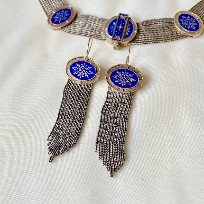 K&ouml;nnte beinhalten: Silberfarbenes Schmuckset mit Halskette und Ohrringen. Die Halskette und die Ohrringe haben ovale blaue Emaille-Akzente mit floralen Designs. Die Ohrringe haben lange, baumelnde Fransen. Das Schmuckst&uuml;ck wird auf einer wei&szlig;en Oberfl&auml;che pr&auml;sentiert.