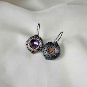 Puede incluir: Un par de pendientes de plata con una piedra preciosa p&uacute;rpura y un dise&ntilde;o de filigrana. Los pendientes est&aacute;n sobre un fondo blanco.