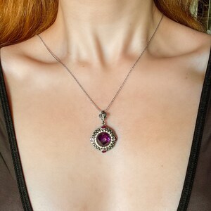 Puede incluir: Un collar de plata con un colgante que presenta una gran piedra preciosa p&uacute;rpura facetada rodeada de piedras m&aacute;s peque&ntilde;as que brillan. El colgante est&aacute; engastado en un marco de plata con un dise&ntilde;o detallado.