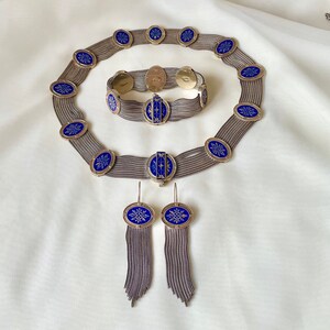 K&ouml;nnte beinhalten: Ein Vintage-Schmuckset mit Halskette, Armband und Ohrringen. Das Set umfasst silberfarbene Metallketten und ovale blaue Emaille-Akzente mit einem wei&szlig;en Schneeflocken-Design. Die Ohrringe haben baumelnde Kettenfransen.