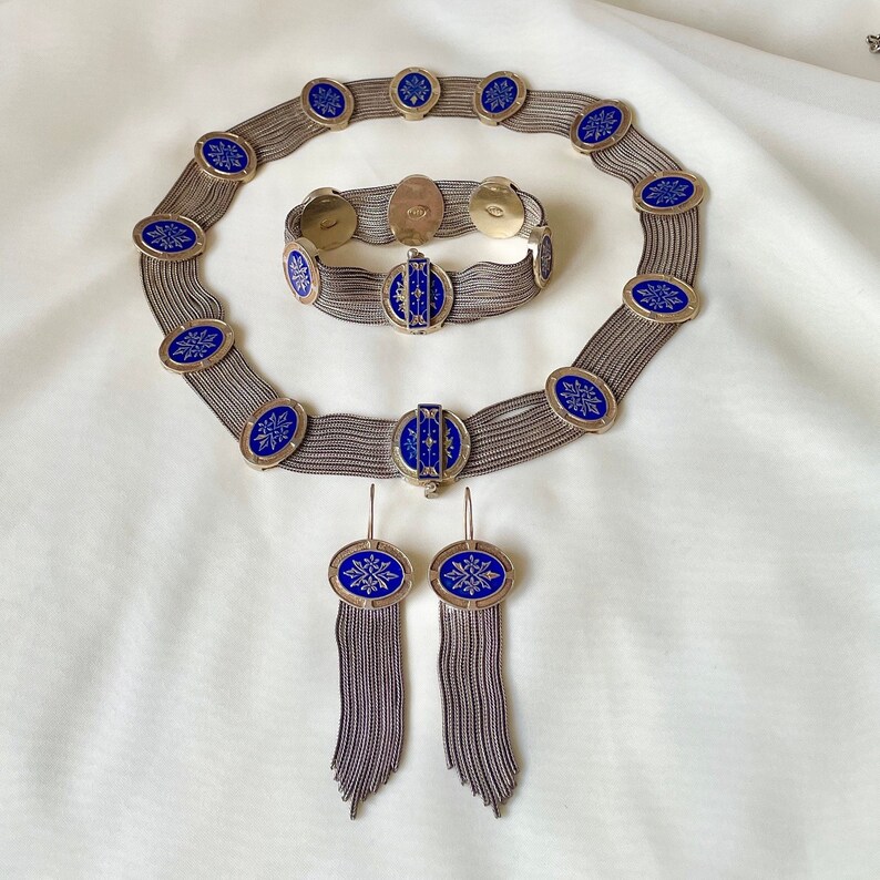 K&ouml;nnte beinhalten: Ein Vintage-Schmuckset mit Halskette, Armband und Ohrringen. Das Set umfasst silberfarbene Metallketten und ovale blaue Emaille-Akzente mit einem wei&szlig;en Schneeflocken-Design. Die Ohrringe haben baumelnde Kettenfransen.