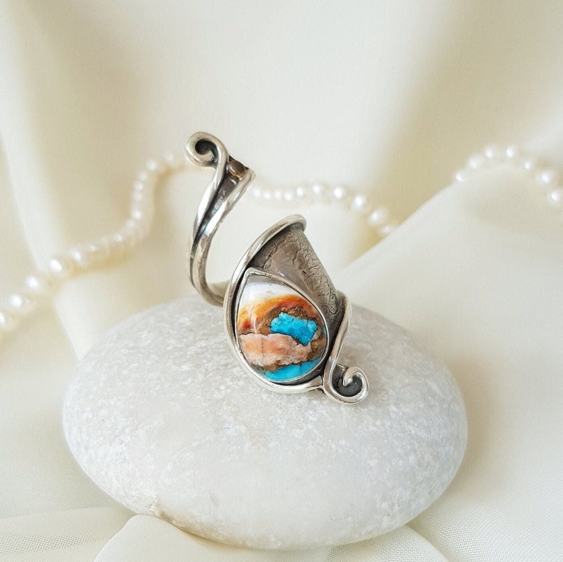 May include: A silver ring with a turquoise and white stone set in a twisted band. The ring is on a white rock.